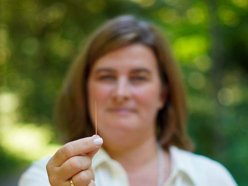 Frau hält eine feine Akupunkturnadel zwischen den Fingern, im Hintergrund grüne Natur