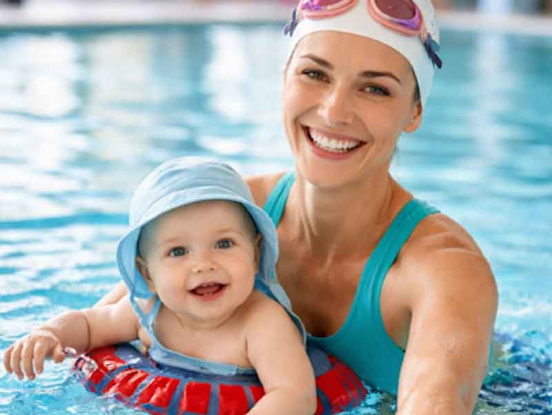 Lachende Mutter mit Badekappe hält ihr Baby mit Schwimmring im Wasser. Erstellt mit KI.