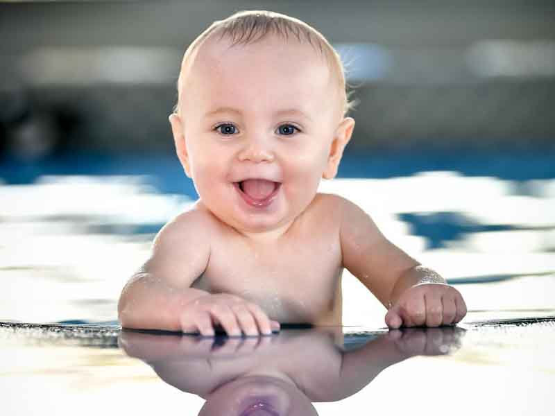 Lachendes Baby taucht strahlend aus dem Wasser auf und stützt sich am Beckenrand ab