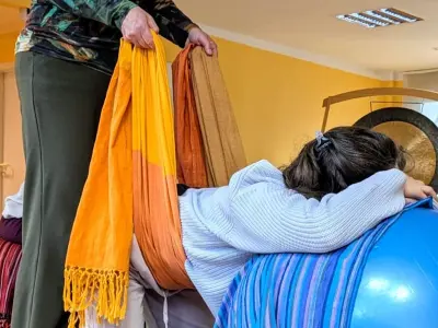 Eine Person lehnt mit dem Oberkörper auf einem blauen Gymnastikball, eine stehende Person hält ein orangefarbenes Rebozo-Tuch an ihrer Hüfte
