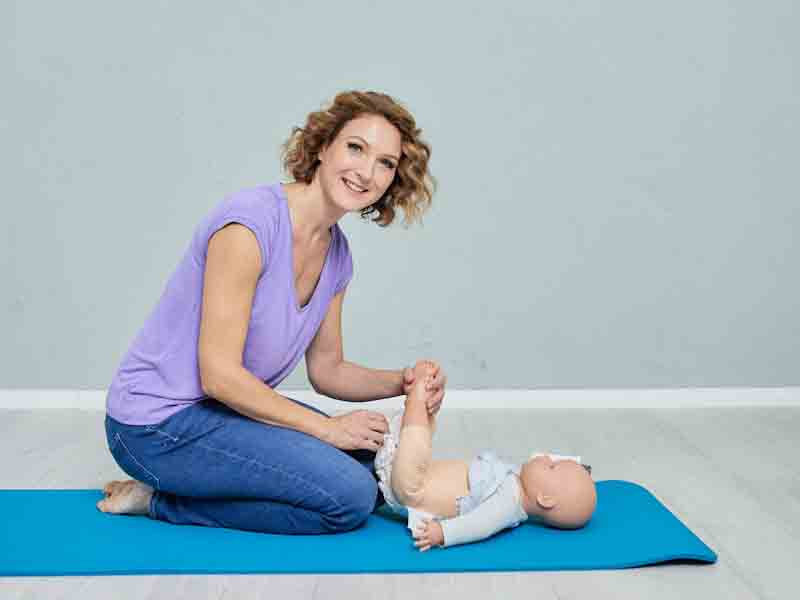 Lächelnde Kursleiterin übt mit einer Baby-Puppe auf einer Matte eine Pflegetechnik